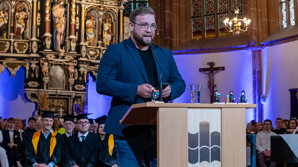 Schulze am Pult, hinter ihm sitzen Professor:innen und Erstsemester.