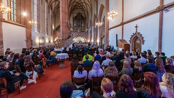 Blick vom Altarraum ins vollbesetzte Kirchenschiff aus dem Altarraum. Zwischen den Köpfen junger Menschen und den Doktorhut-geschmückten Köpfen von Professor:innen steht eine große Erntekrone.