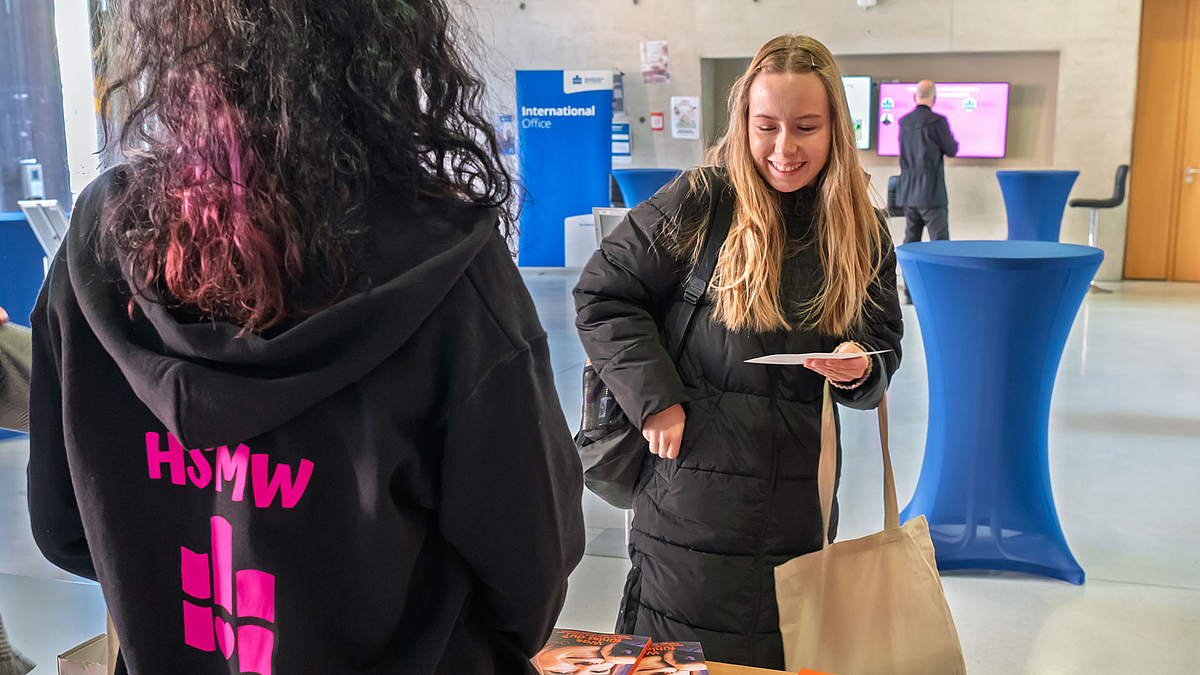 Eine junge weibliche Person steht lächelnd vor einem Tisch. Ihr gegebüber im Bild von hinten zu sehen steht eine Person im schwarzen Hoodie mit pinken Rückenprint aus HSMW-Schriftzug und HSMW-Logo.