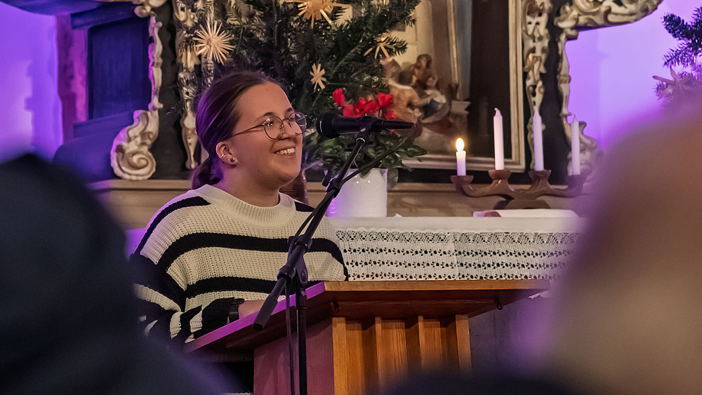 Eine junge weibliche Person steht lächelnd am Pult vor dem Altar uns spricht in ein Mikrofon.