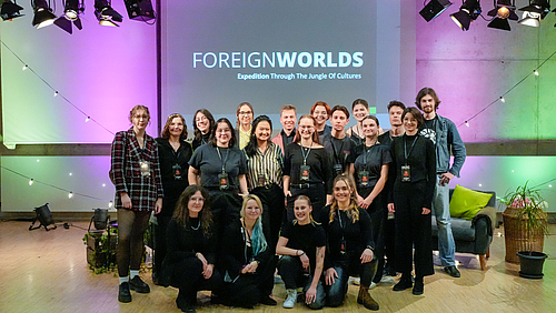 Gruppenbild von 19 jungen Personen in einer dekorierten Studioatmosphäre, von den einige knien. Im Hintergrund ist auf einer Leinwand der Schriftzug „Foreign Worlds. Expedition Through The Jungle of Cultures“ zu lesen. 