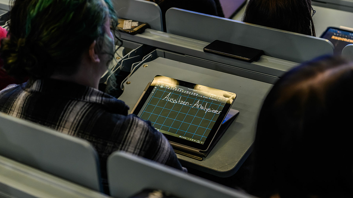 Blicke über die Köpfe auf ein Tablet auf den Klapptisch eines Platzes im Hörsaal. Auf dem Display ist die Handschriftnotiz „Aussitzen – Analysieren“ zu sehen.