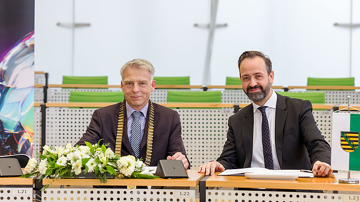 Zwei männliche Personen mit Anzug und Krawatte sitzen an Abgeordnetenbänken eines Plenarsaals. Die linke Person trägt eine goldene Amtskette. Vor ihnen liegen aufgeschlagene Dokumente, die auf der linken Seite von einem Blumengesteck zum größten Teil verdeckt werden. Am rechten Bildrand ist ein Tischbanner mit dem sächsischen Wappen zu sehen.