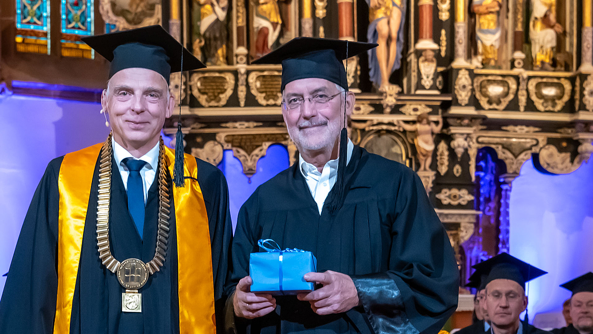 Rechts neben dem Rektor steht der emeritierte Professor Christian Schulz, Verfalls in Talar und Doktorhut. Er hält ein kleines Geschenkpäckchen in der Hand.