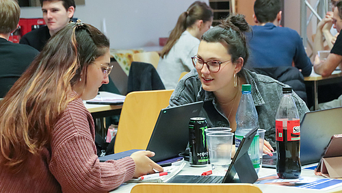 Gute Strategie: engagiert und gut versorgt an der Hochschule Mittweida. Zwei junge weibliche Personen an einem Tisch mit Laptops, Bechern, Colaflaschen und Energydrinks in Dosen. Die Frau rechts wendet sich der linken interessiert zu. Im Hintergrund sind weitere Personen an Tischen zu sehen.