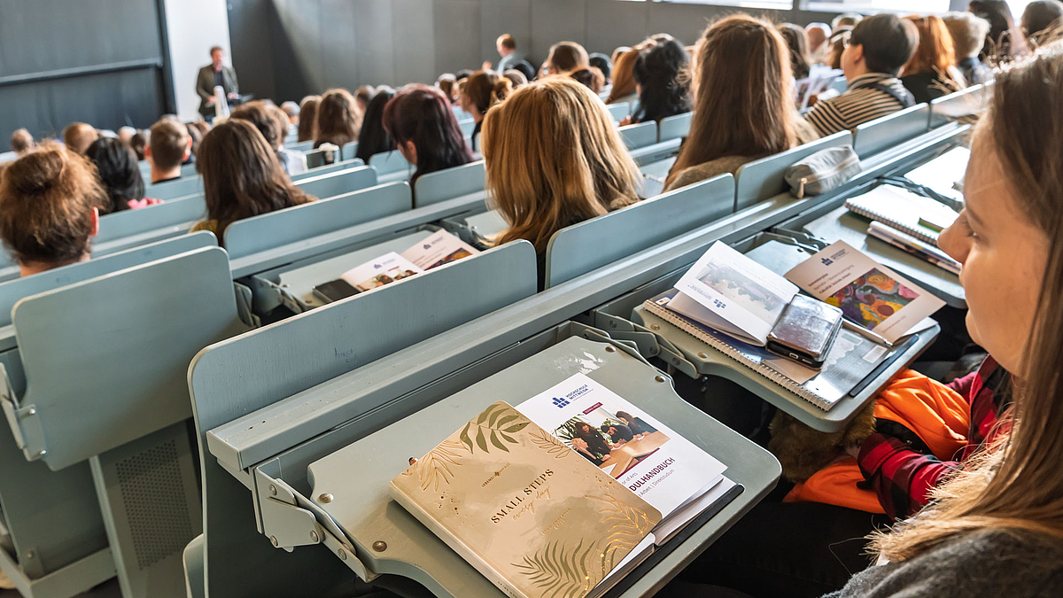 Blick über dei Schulteren einer Person auf deren Bank im Hörsaal. Vor ihr auf dem Klapptisch liegen ein Notitbuch mit dem Title "Small Steps every Day" und ein Modulhandbuch.