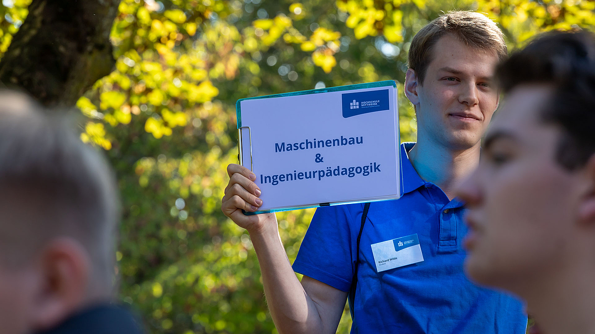 Ein junger Mann hält ein Schild mit der Aufschrift "Maschinenbau & Ingenieurpädagogik".