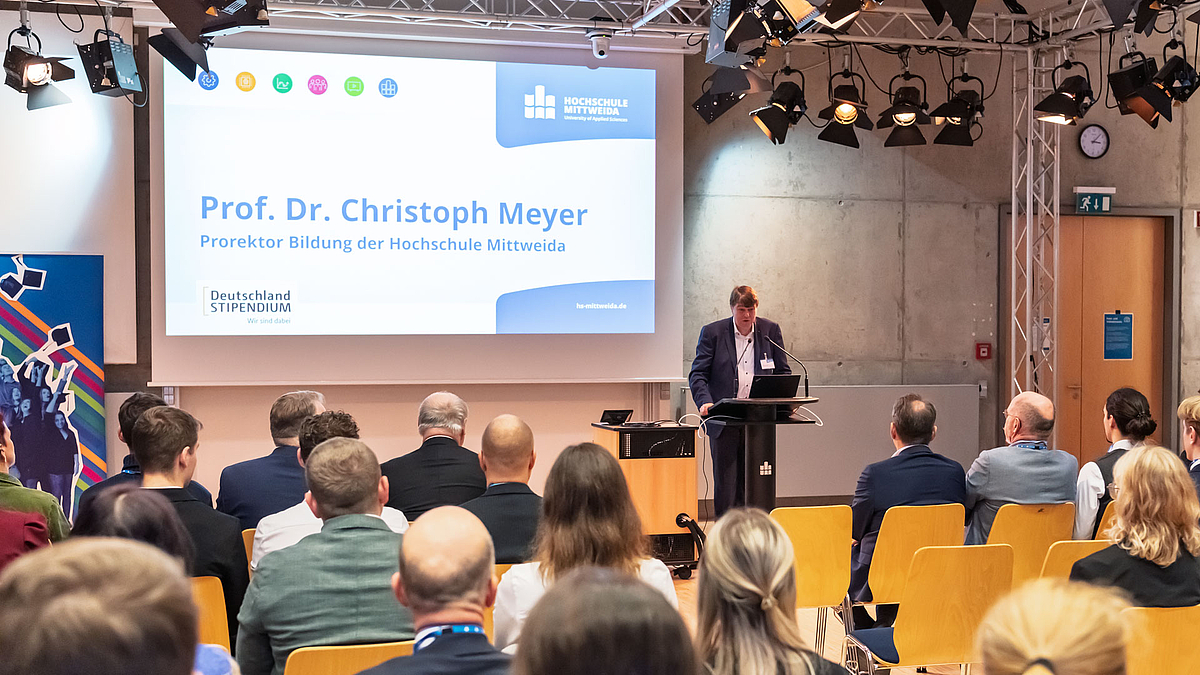 Eine männliche Person im Anzug ohne Krawatte spricht von einem schwarzen Pult aus zum Publikum, das von hinten zu sehen ist. Auf der Leinwand hinter ihm sind sein Name und seine Funktion projiziert sowie die Logos der Hochschule und des Deutschlandstipendiums.