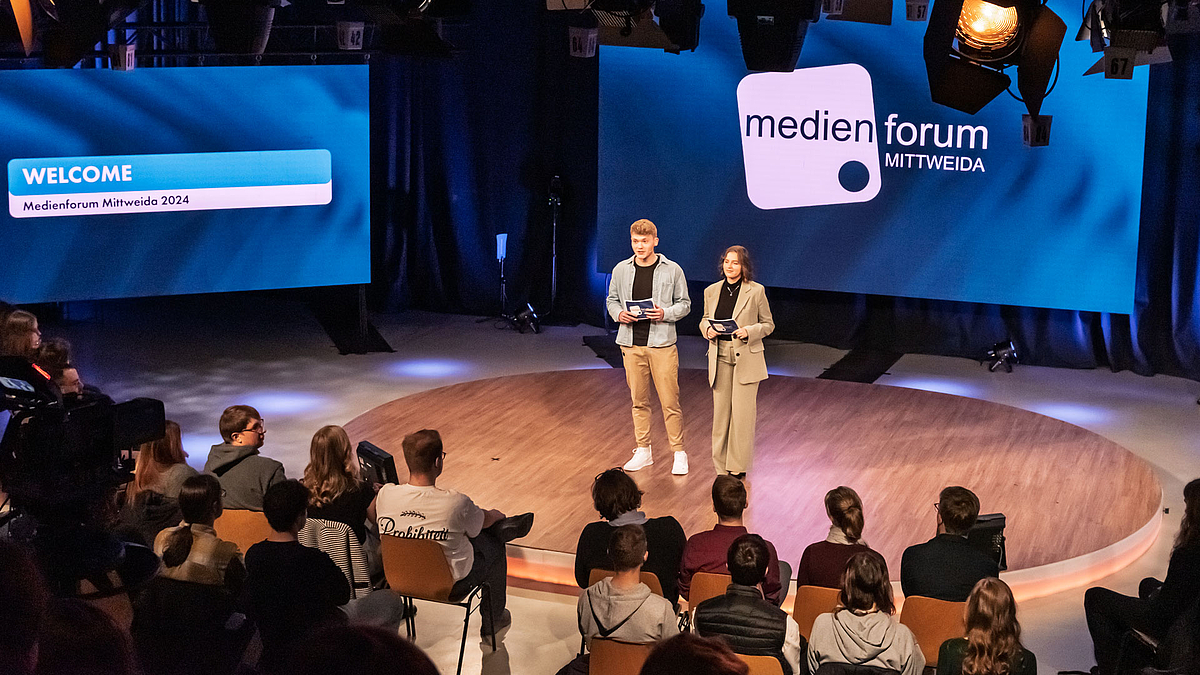 Eine junge Mann und eine junge Frau im Fernsehstudio sprechen zum Publikum im Vordergrund. Auf den Screens ist das gleiche zu sehen wie auf dem Foto zuvor.