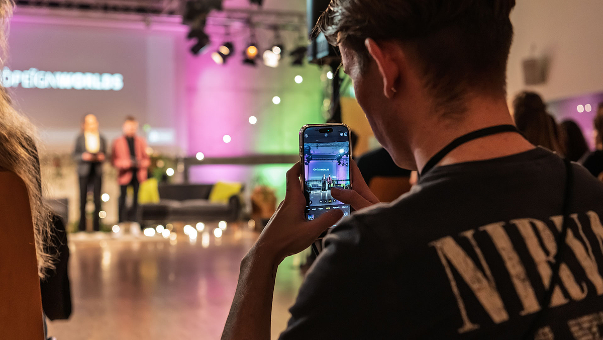 Blick durch das Display eines Smartphones, das gerade die Moderator:innen aufnimmt, die im Bildhintergrund unscharf zu erkennen sind.