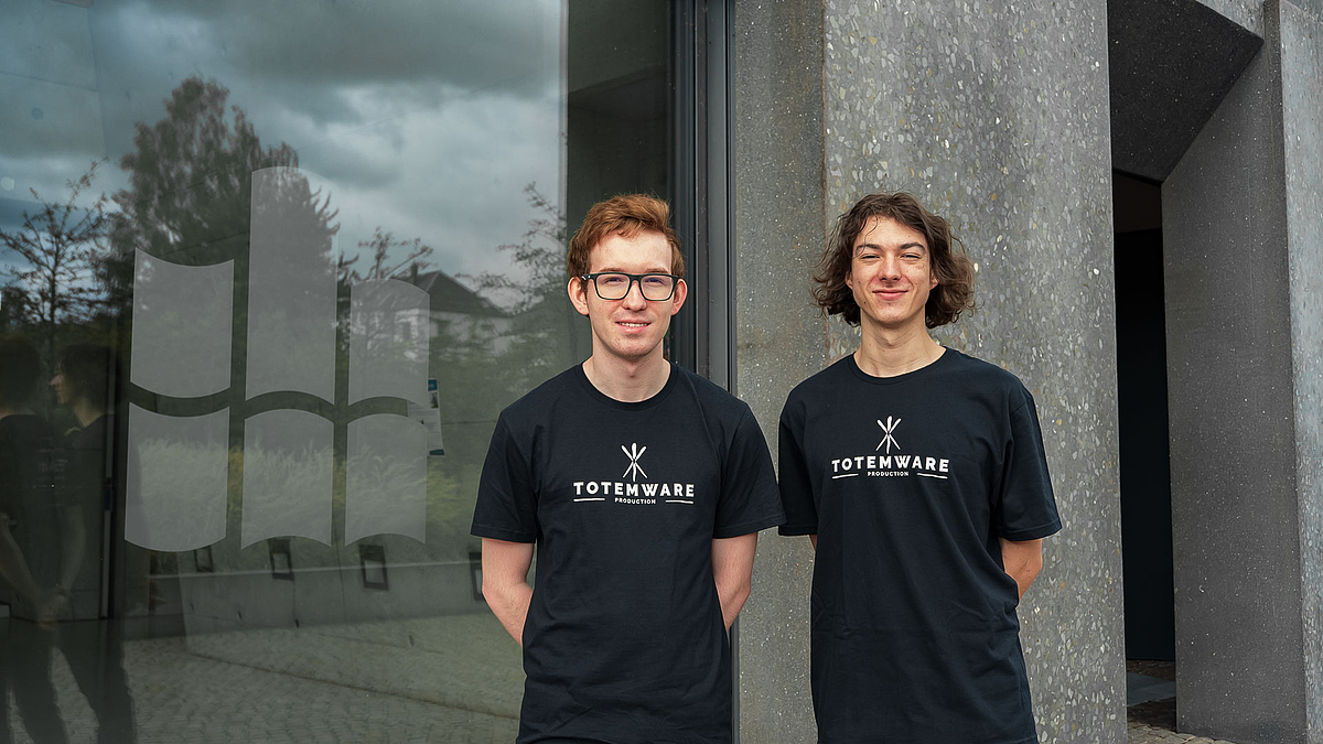 AT: Zwei Studenten stehen vor vor einem Fenster mit HSMW-Logo, ihre T-Shirts tragen Logo und Schriftzug „TOTEMWARE.“