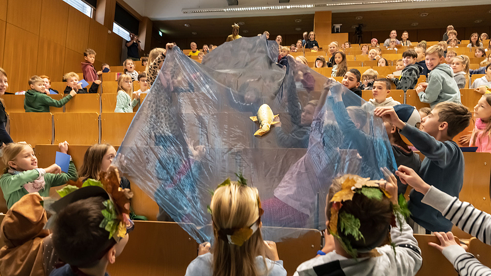 Kinder im Hörsaal haben eine blaue Folie von oben nach unten aufgespannt und transportieren mit geschickten Bewegungen der Folie einen Plastikfisch über ihre Köpfe nach oben.