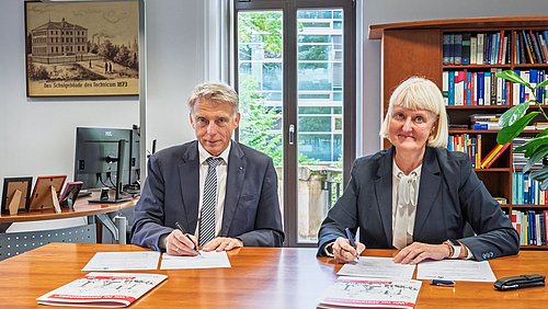 Rektor Volker Tolkmitt und die Vorsitzende der Geschäftsführung der Freiberger Arbeitsagentur Kathrin Groschwald besiegeln die engere Kooperation für ein verbessertes Orientierungs- und Beratungsangebot. Eine männliche und eine weibliche Person sitzen nebeneienander an einem Tisch und blicken in die Kamera. Vor ihnen liegen jeweils Dokumente, die sie soeben unterschreiben haben.