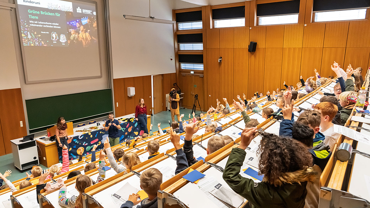 Die Bankreihen im Hörsaal sind voller Kindern, die zum Melden ihre Finger nach oben strecken. Unten vor dem Pult stehen die beiden Dozentinnen, rechts ein Kameramann im Tierkostüm. Zwischen den beiden Dozentinnen steht eine männliche Person mit Handmikrofon und spricht zu den Kindern. Auf der Leinwand im Hintergrund ist „Kinderuni. Grüne Brücken für Tiere“ zu lesen.