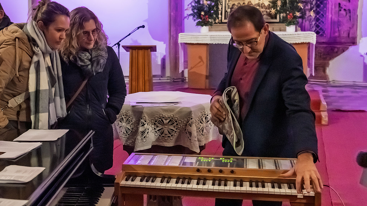 Zwei weibliche Personen schauen interessiert auf eine Tastatur, die im rechten Winkel zur Tatstatur eines Flügels aufgebaut ist und einem elektronischen Keyboard ähnelt . Professor Czerner beugt sich über den Spieltisch und betätigt eine Taste.