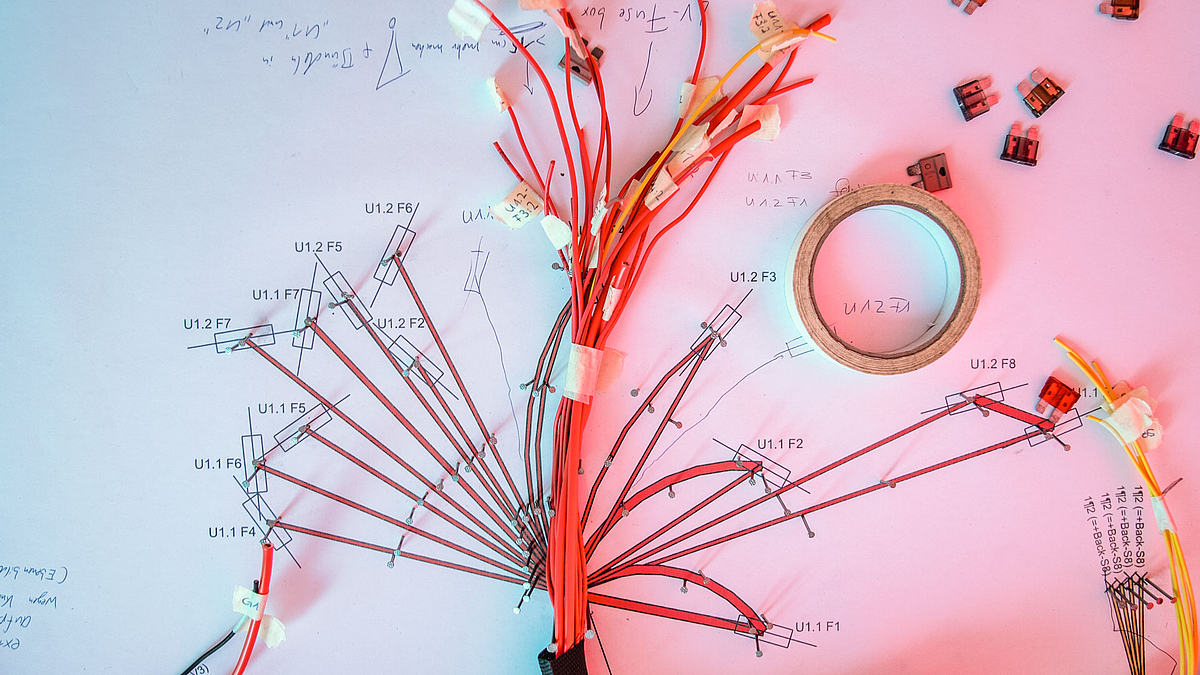 Zahlreiche Kabel aus einem Kabelschlauch verzweigen sich auf einem beschrifteten Untergrund und sind mit Nägeln darauf befestigt.