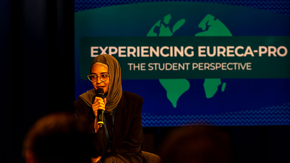 Eine Studentin mit Kopftuch im Fernsehstudio. Sie spricht in ein Handmikrofon. Hinter ihr auf einem Monitor ist zu lesen: "Experiencing EURECA PRO. The Student Perspective"