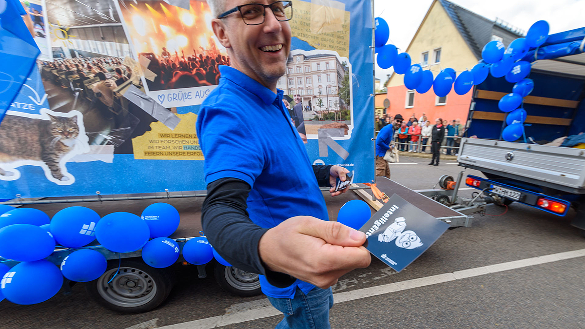 Ein Mitarbeiter der Hochschule im blauen T-Shirt läuft vor einem Motivwagen. Er hält einen Aufkleber mit dem Motiv einer Ente und der Unterschirft "IntelligEnte" in die Kamera.