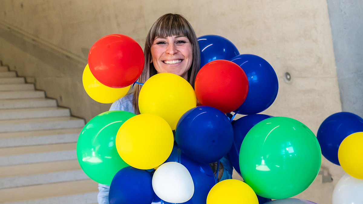 Eine junge weibliche Perso schaut strahlden hinter bunten Luftballons hervor, die sie vor sich hält.