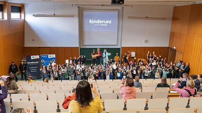 Hörsaal mit ansteigenden Bankreihen. Unten stehen rund einhundert fröhliche Kinder mit Urkunden. Hinter ihnen und Teil auf dem Pult stehe die Mitglieder des Kinderuni-Teams in ihren Tier-Kostümen. In den Bankreihen stehen Eltern, die mit ihren Smartphones Fotos machen.