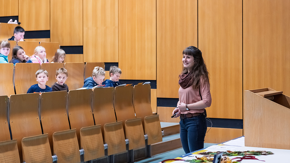 Eine junge Frau mit Kopfbügelmikrofon steht lächelnd zwischen Pult und Bankreihen des Hörsaals. Im Hintergrund sind Kinder in einer Bankreihe zu sehen.