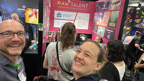 Selfie bei der Raw Talent-Premiere: Alexander Marbach mit seiner Kölner Professor:innen-Kollegin Greta Hoffmann. Zwei Menschen schauen freundlich grinsend in die Kamera, während sie ein Selfie machen. Im Hintergrund steht ein junger Mann und spielt an einem Messestand ein Computerspiel.