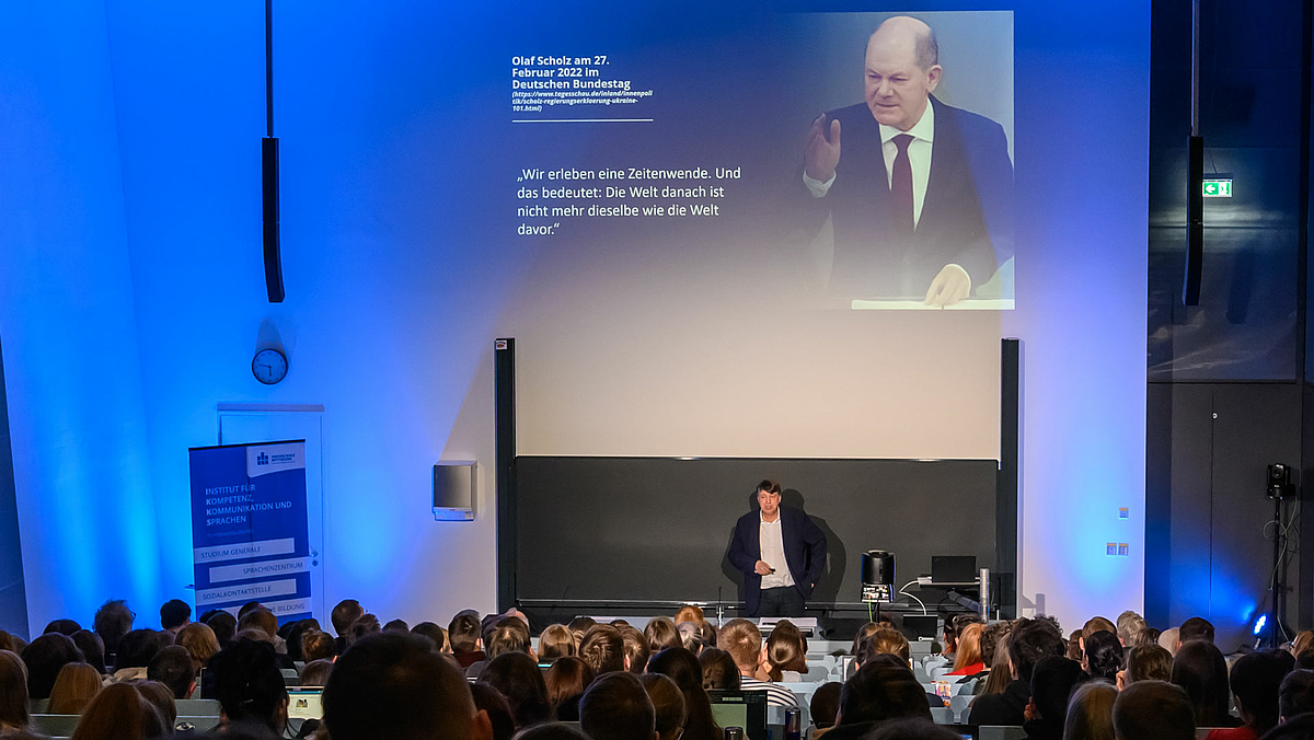 Blick von hinten über die Köpfe der Zuschauenden im voll besetzten Hörsaal auf den männlichen Dozenten, der vor der Tafel steht. Über ihm zeigt die Projektion rechts ein Bild von Bundeskanzler Olaf Scholz bei einer Rede und links das Zitat aus der Regierungserklärung vom 27. Februar 2022: „Wir erleben eine ‚Zeitenwende‘. Und das bedeutet: Die Welt danach ist nicht mehr dieselbe wie die Welt davor.“