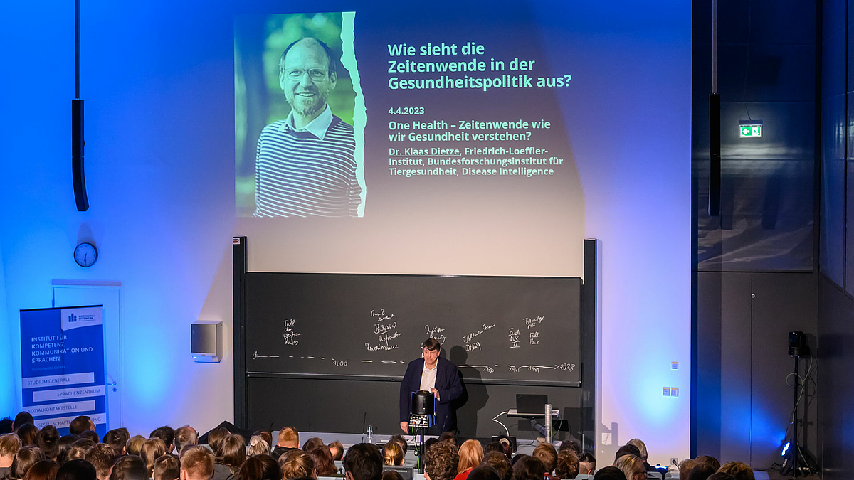 Blick von hinten über die Köpfe der Zuschauenden auf den Dozenten, der vor der Tafel steht. Über ihm zeigt die Projektion links das Portrait einer männlichen Peron und rechts die Worte: „ Wie sieht die Zeitenwende in der Gesundheitspolitik aus. 4.4.2023. One Health – Zeitenwende wie wir Gesundheit verstehen? Dr. Klaas Dietze, Friedrich-Loeffler-Institut für Tiergesundheit. Desease Intelligence.“