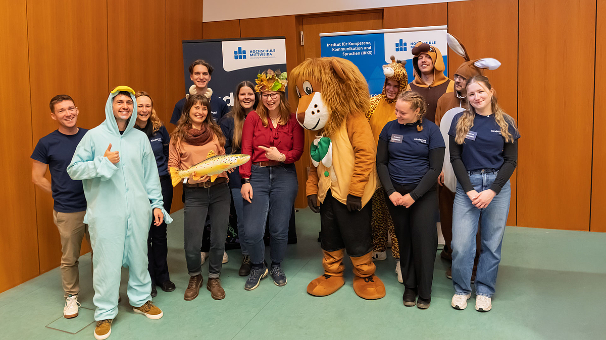 Sechs männliche Personen, zum Teil in Tier-Kostümen, und sechs weibliche Personen stehen im Hörsaal fröhlich lachend vor zwei Rollups mit Logos und Schriftzügen „Hochschule Mittweida“ und „Institut für Kompetenz, Kommunikation und Sprachen (IKKS)“. Zwischen den Personen steht das Stadtmaskottchen Mitti. Eine Person hält eine Plastik-Forelle in den Händen.