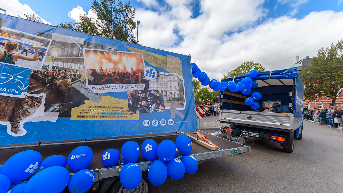 Sichtbar ist ein kleiner LKW, der mit einer Girlande mit blauen Ballons mit einem Motiv-Anhänger versehen ist. Auf der Motiv-Plane sieht man unter anderem eine "Campuskatze", eine Sportlerin, einen vollen Hörsaal, einen Schwarzen Studenten mit Absolventenhut und eine Konzertbühne vor der Menschen tanzen.