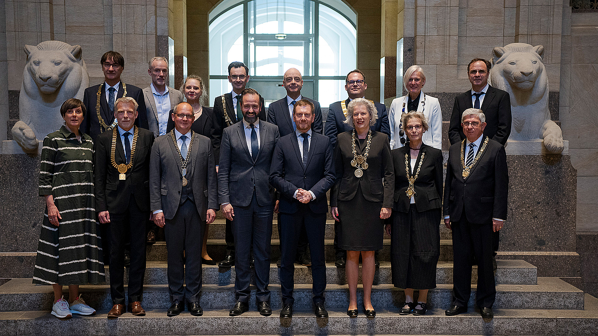 14 Männer und Frauen stehen gemeinsam mit Wissenschaftsminister und Ministerpräsident auf einer Steintreppe. Im Hintergrund stehen zwei Löwenplastiken aus hellem Stein.