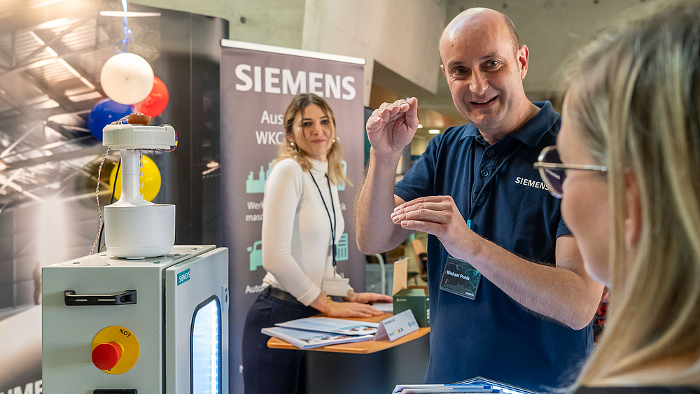 Ein Vertreter der Firma Siemens im Gespräch mit einer Standbesucherin. Im Hintergrund ist eine weitere Vertreterin von Siemens zu sehen, die in Richtung Kamera blickt.