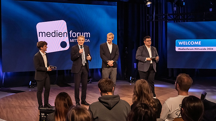 Vier männliche Personen stehen auf einem leicht erhöhten Podest im Fernsehstudio. Im Vordegrund sitzen Zuschauer:innen. Auf den Screens im Hintergrund sind das Medienforum-Mittweida-Logo und der Schriftzug "WELCOME. Medienforum Mittweida 2024" zu sehen.. 