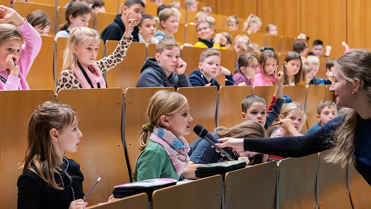 Ein Mädchen in einer Bankreihe spricht in ein Mikrofon, das eine Kinderuni-Mitarbeiterin ihr hinhält.