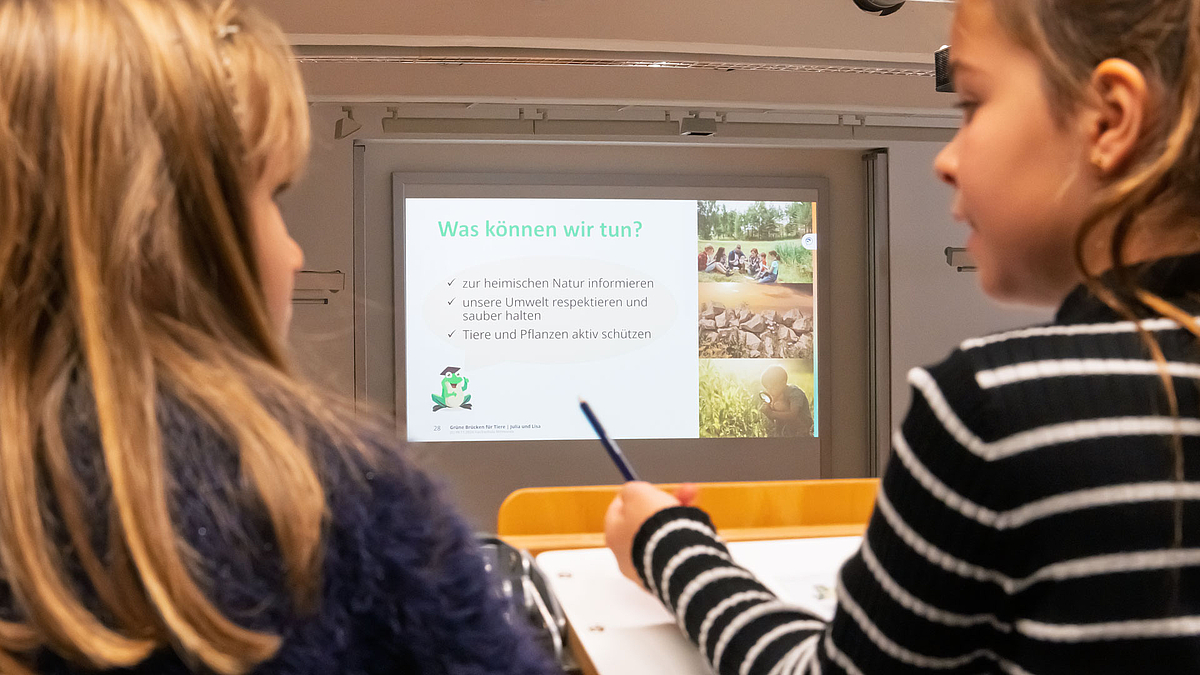 Blick zwischen zwei Mädchen hindurch auf die Leinwand mit dem Text: „Was können wir tun?“ und den drei Anstrichen: „zur heimischen Natur informieren; unsere Umwelt respektieren und sauber halten; Tiere und Pflanzen aktiv schützen“.