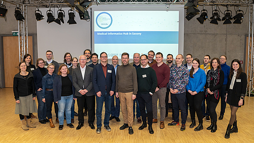 Gruppenaufnahme mit 26 Personen. In Hintergrund ist eine Folie projiziert mit dem MiHUBx-Logo und dem Schriftzug „Medical Informatics Hub in Saxony“.