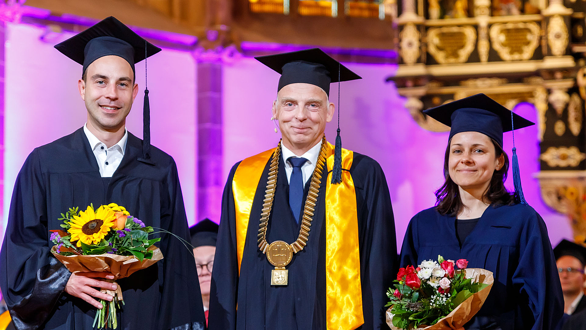 Rektor Volker Tolkmitt steht im Altarraum zwischen einem Mann und einer Frau. Alle drei tragen Talar und Doktorhut, der Rektor zusätzlich seine goldene Amtskette. Die beiden jungen Professor:innen haben Blumensträuße in der Hand.