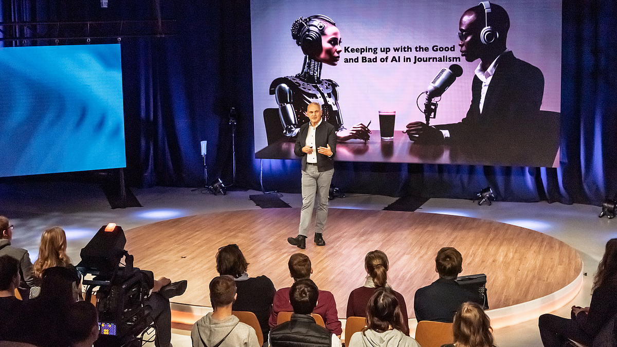 Ein Mann im Studio spricht zum Publikum vor einem Screen, der einen Tisch zeigt, an dem ein Roboter im Gespräch mit einem Menschen ist. Zwischen den beiden steht der Text:„Keeping  up with the Good and the Bad of AI in Journalism“.