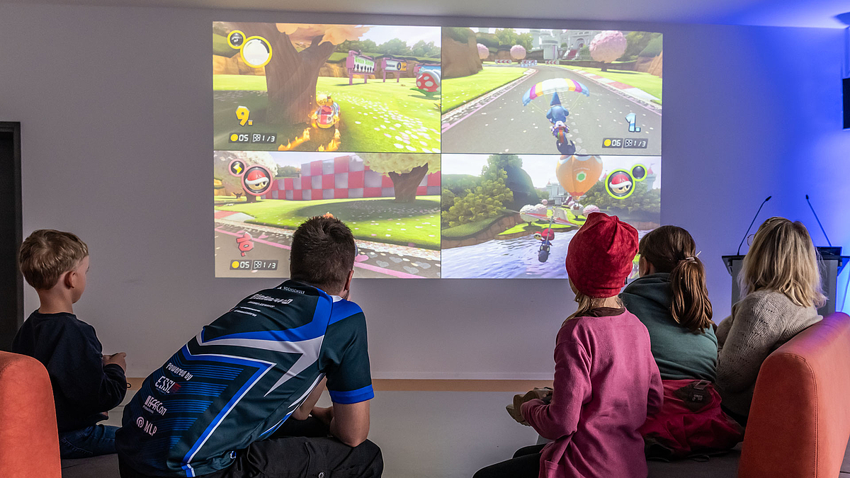 Auf einer Leinwand sieht man das Spiel Mario Kart - im Vordergrund sitzen fünf Menschen, vier davon spielen das Spiel gegeneinander.