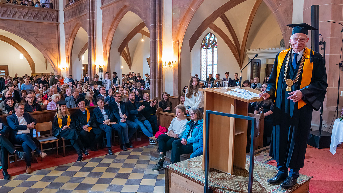 Blick vom Altarraum ins Kirchenschiff. Einige Menschen stehen. Am rechten Bildrand am Pult stehend wendet sich der Rektor lächelnd nach hinten.