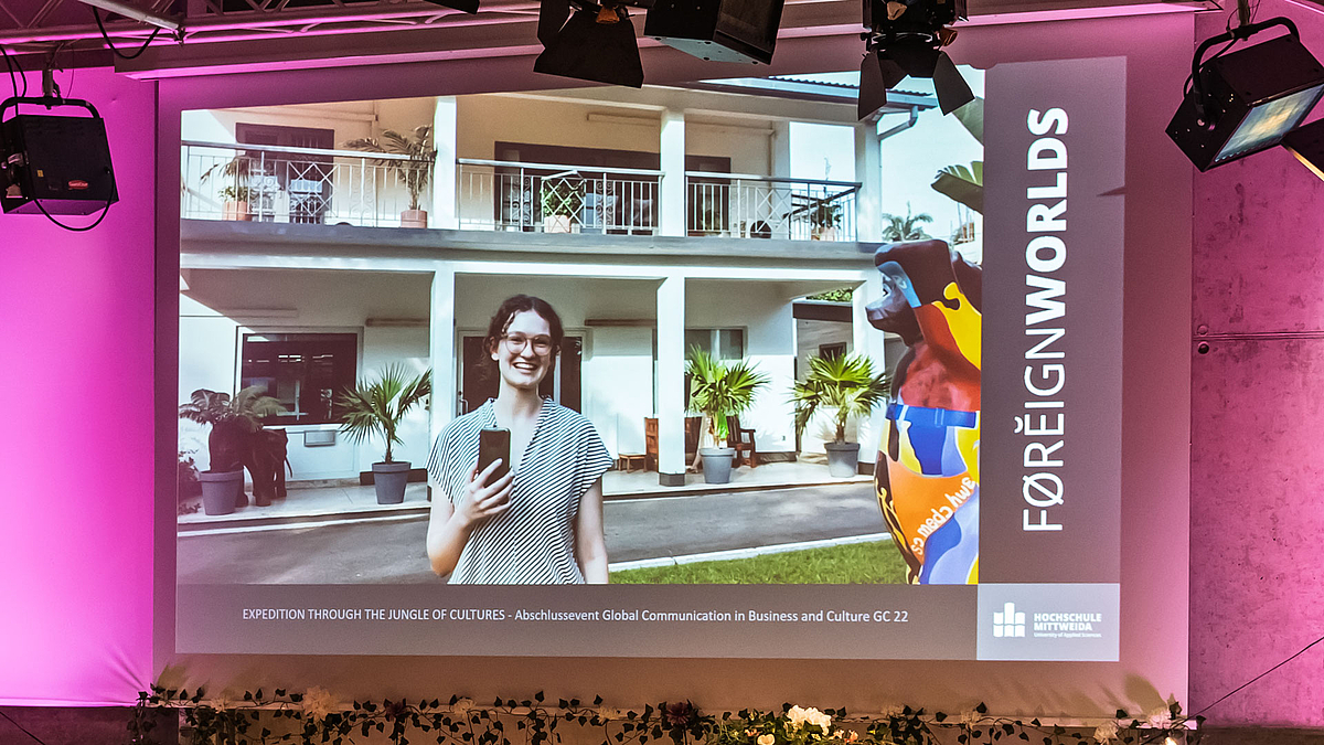 Auf der Leinwand ist ein Video zu sehen, in dem eine junge weibliche Person vor einem Gebäude mit Säulen und Palmen in Richtung Kamera spricht.