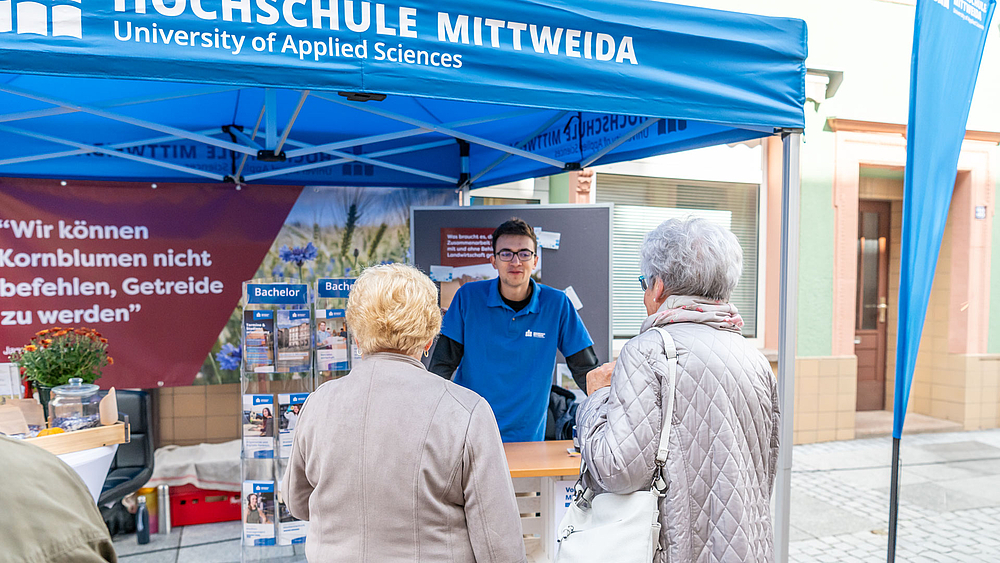 Zwei ältere Frauen mit weißen Haaten stehen vor einem jungen Mann. Es ist ein Flyerständer zu sehen und ein offenes Zelt mit der Aufschrift "Hochschule Mittweida".