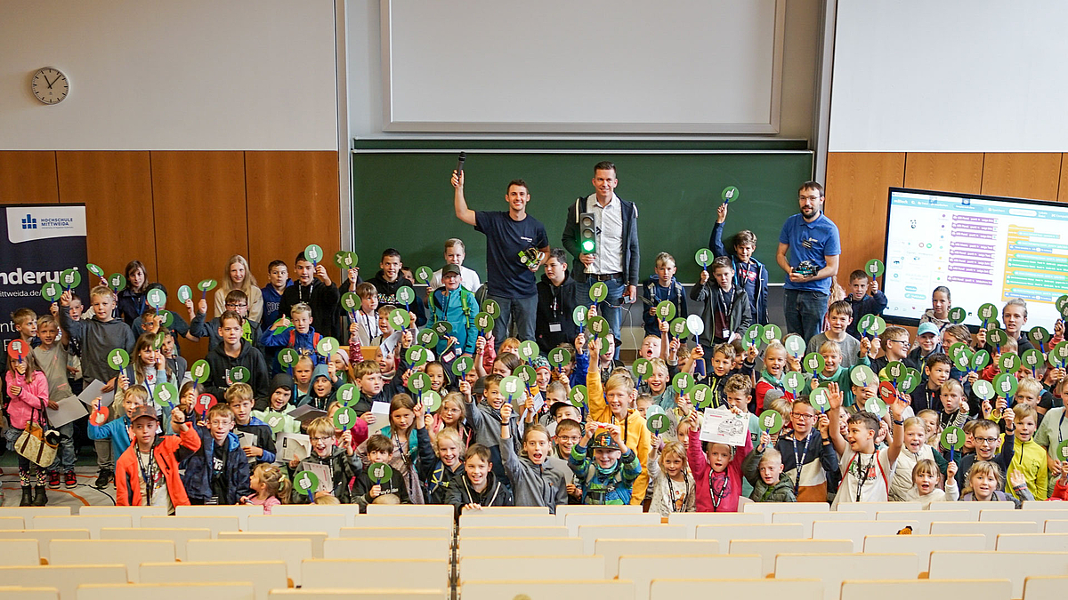 Viele Kinder und ihre Dozenten stehen am unteren Ende des Hörsaals und winken fröhlich mit „Daumen-hoch“-Schildern. Hinter ihnen auf dem Pult stehen der Erwachsene.