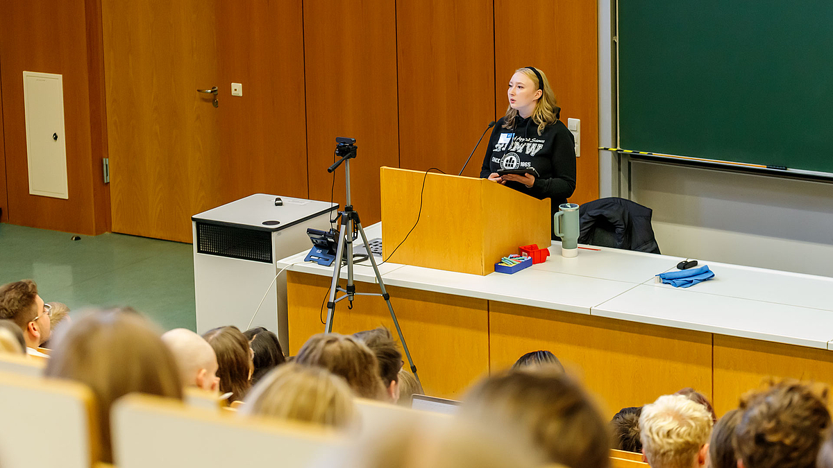 Blick von oben über die Köpfe von Zuhörenden im Hörsaal hinweg auf eine junge Frau am Pult.