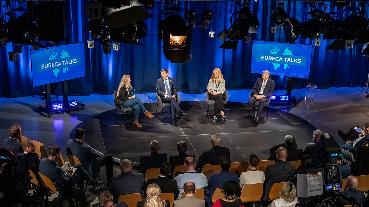 Vier Personne auf einem Podieum im Fernsehstudio. Im Vordergrund sind Zuschauer:innen von hinten zu erkennen.