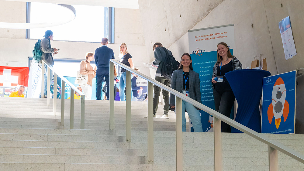 Blick von unten auf die große Treppe im Ludwig-Hilmer-Bau mit dem Stand des Gründernetzwerks Saxeed auf dem Zwischenpodest. Zwei junge Frauen vom Stand lachen in Richtung Kamera.